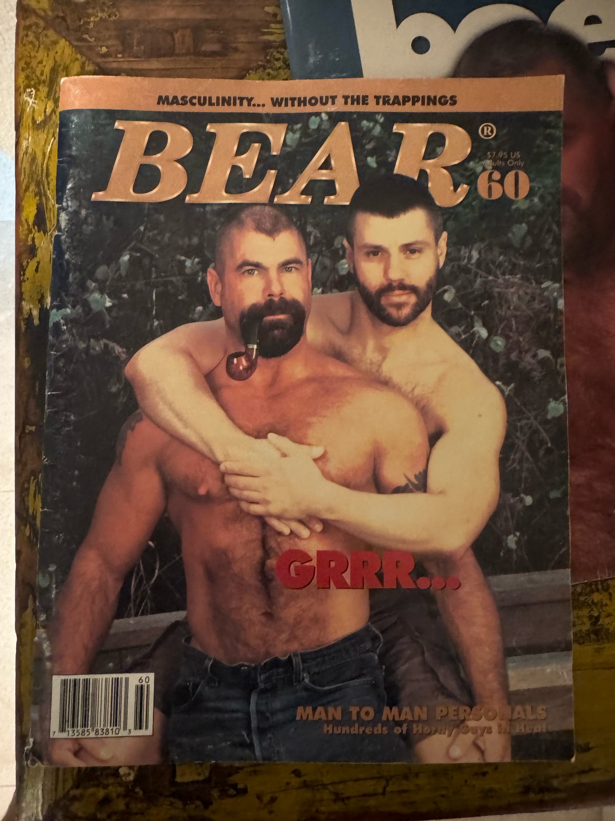 A worn cover of a vintage bear-community magazine showing two bare-chested men embracing, photographed as a keepsake; colour.