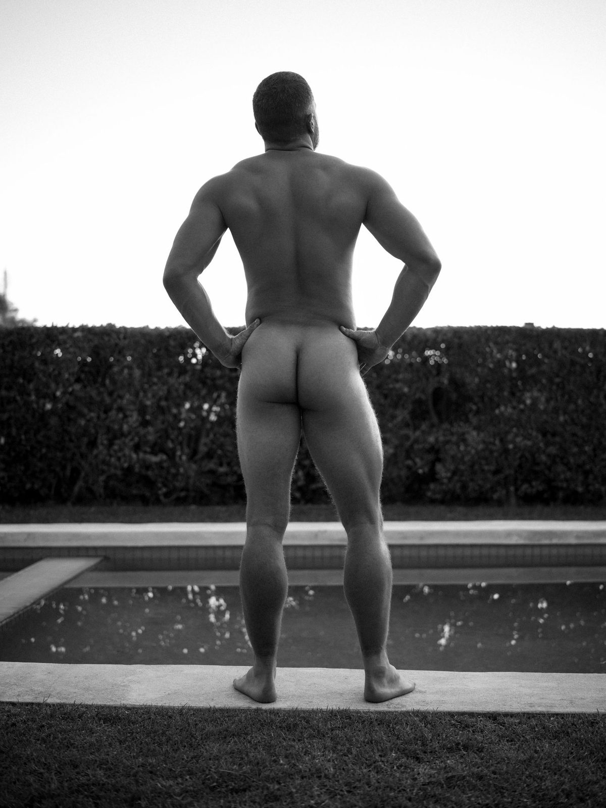 A muscular nude man stands by a poolside lawn at dusk, seen from behind with hands on his hips, looking out; black and white.