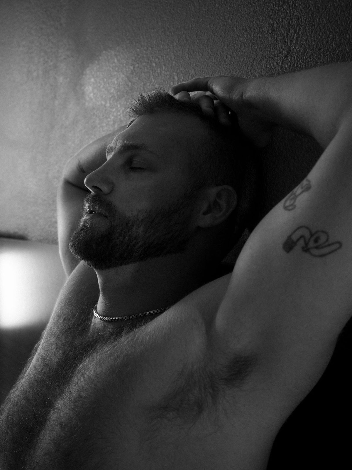 A bearded man, bare-chested, reclines with arms raised behind his head and eyes closed, shot close and intimate; black and white.