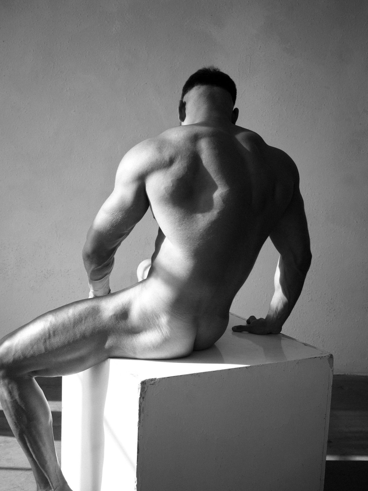 A muscular nude man sits on a white pedestal, seen from behind, glancing back over his shoulder; black and white.