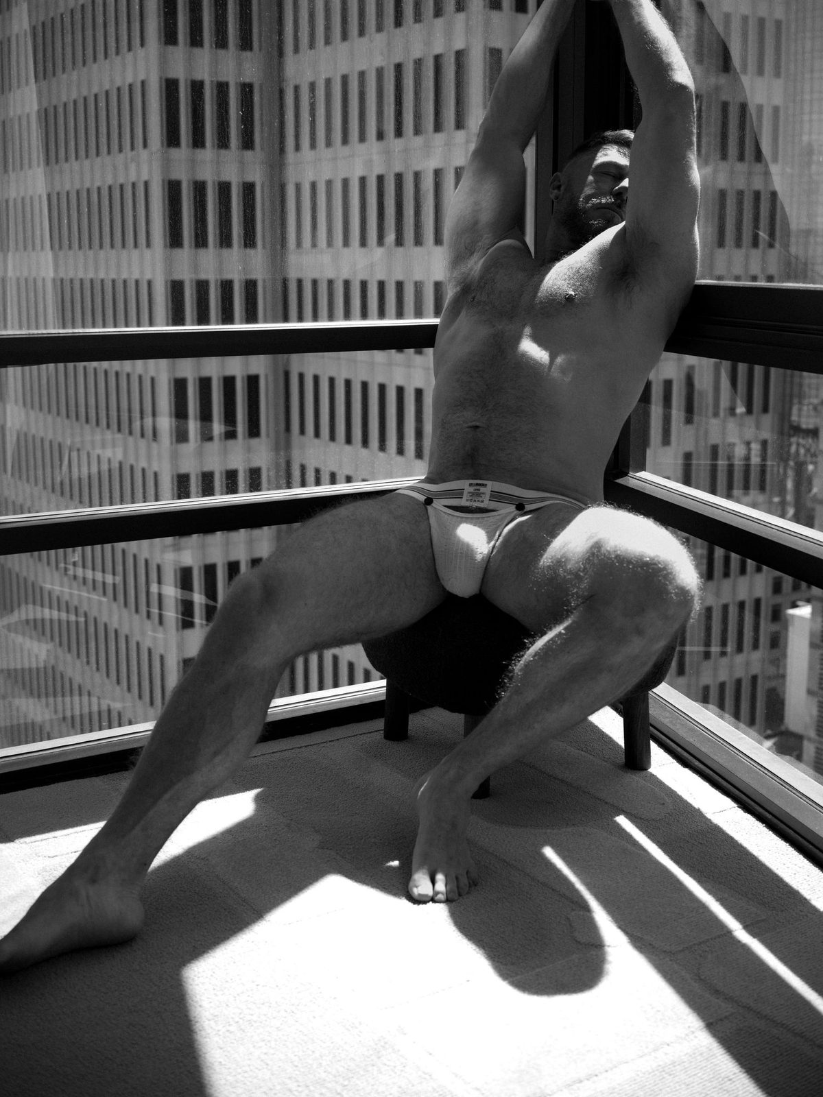 A man in a jockstrap crouches on a sunlit high-rise balcony, arms stretched overhead, skyline behind him; black and white.