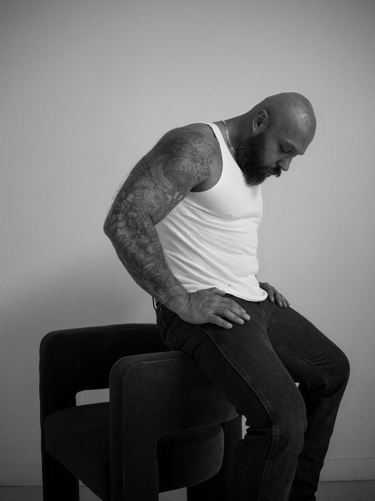 A bald, bearded, tattooed man in a white tank top sits astride a chair, looking down, arms resting on its back; black and white.