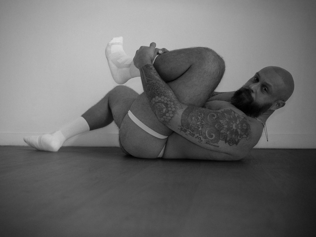 A bearded, tattooed man lies on a studio floor with one knee drawn up in his arms, looking back at the camera; black and white.