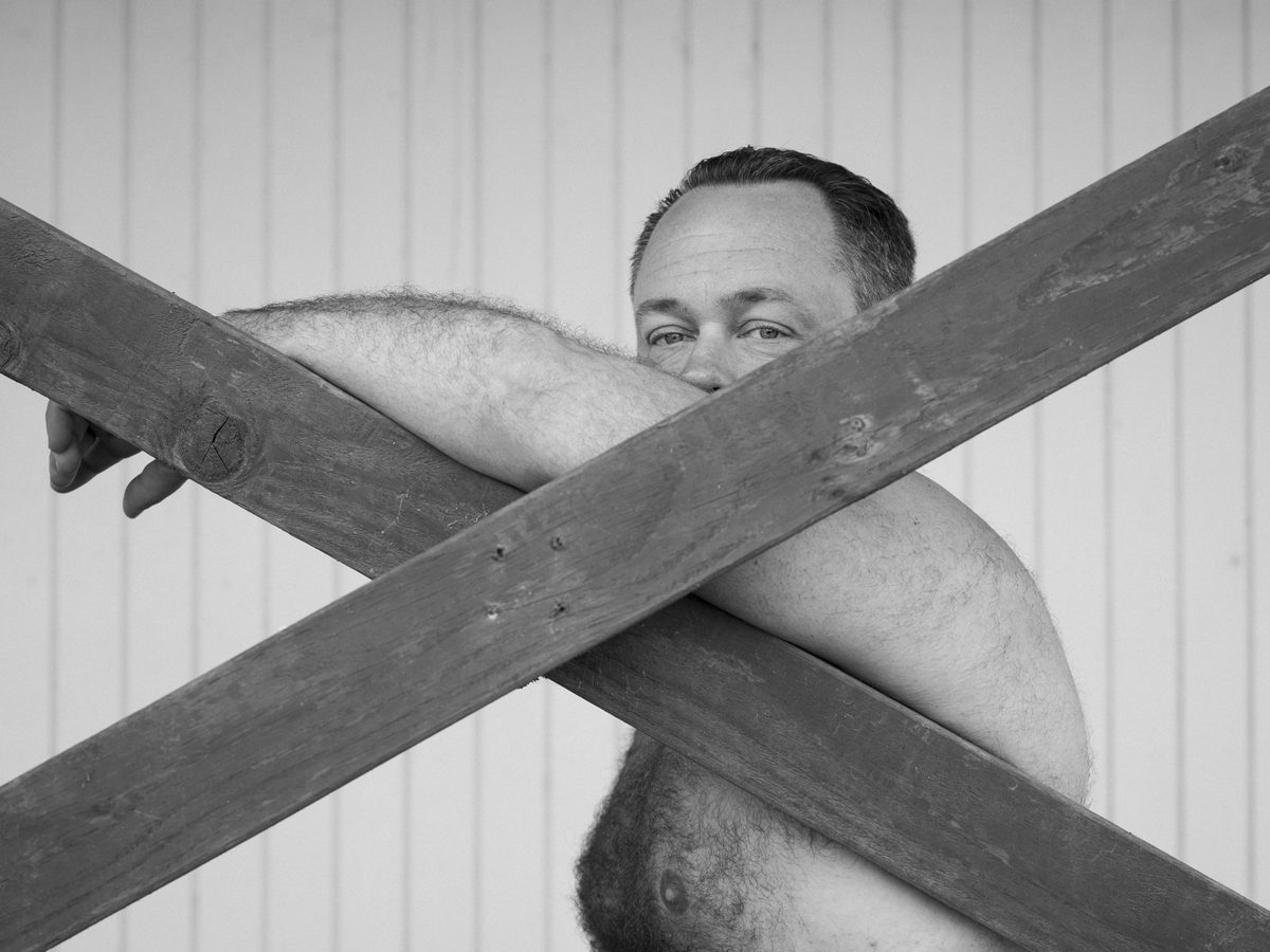 Spread 1 of 7 from Lazy Bear: a bearded man leans against crossed wooden beams, bare-chested, looking at the camera; black and white.