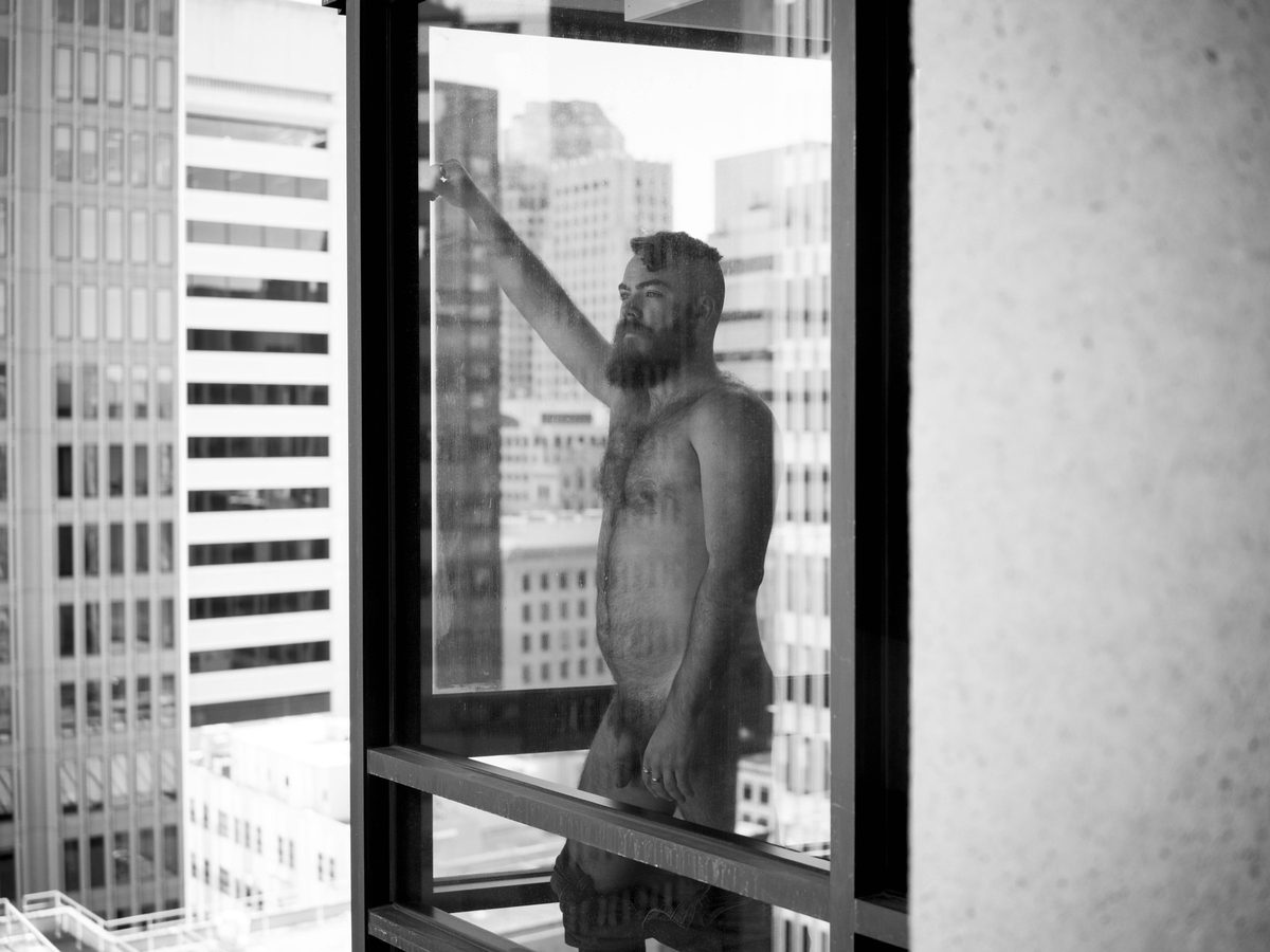 Spread 1 of 7: a bearded man stands nude at a high-rise window, one arm raised to the glass, city towers beyond; black and white.