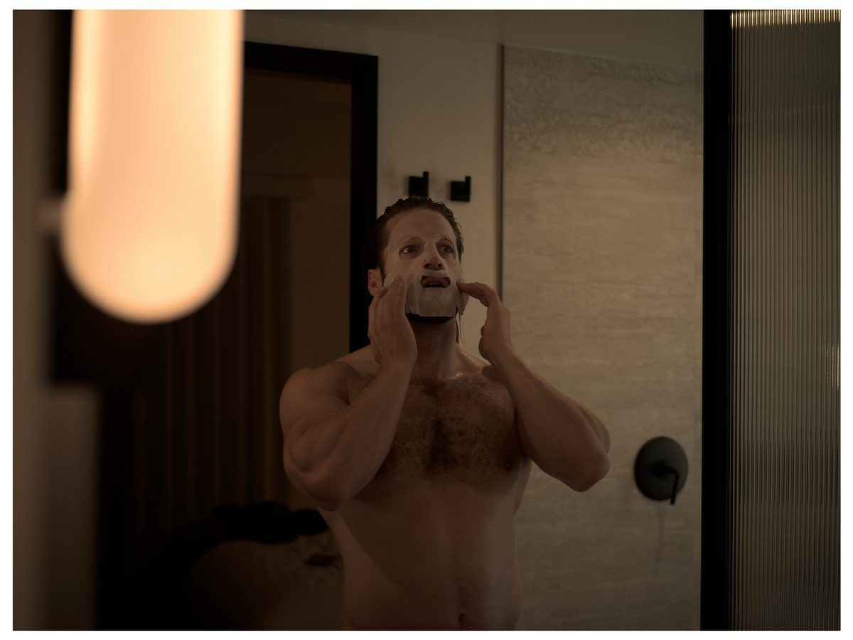 Spread 11 of 15 from An American Pyscho: a muscular, bearded man applies a white face mask at a bathroom mirror; colour, low light.