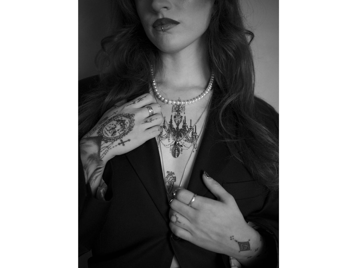 Spread 3 of 7 from The Apprentice: a close portrait of a tattooed woman in a black blazer, hand at her chest, wearing layered necklaces; black and white.