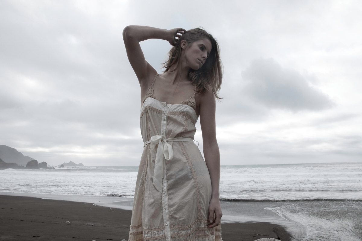 Spread 6 of 7 from The Apprentice: a woman in a pale slip dress stands on a dark sand beach, wind in her hair; colour, overcast.