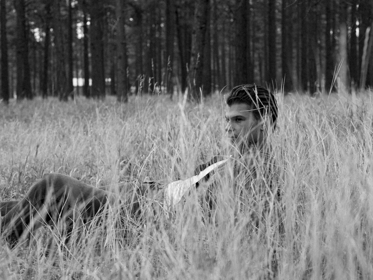Spread 1 of 6 from The Woodsman: a young man crouches in tall grass at a forest edge, resting a hatchet, looking aside; black and white.