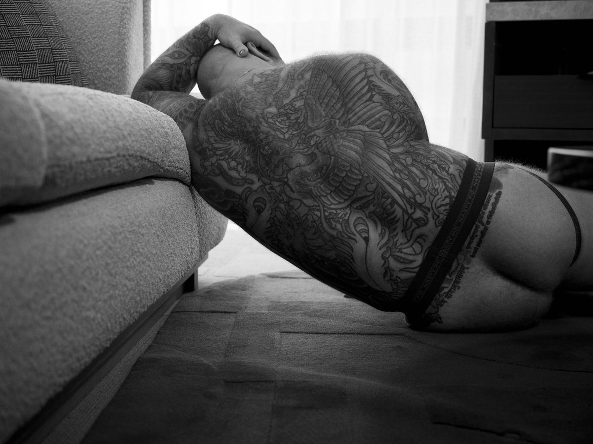Spread 2 of 8 from Beef, Brawn and Bare: a heavily tattooed man lies across a sofa in briefs, seen from behind, arm raised behind his head; black and white.