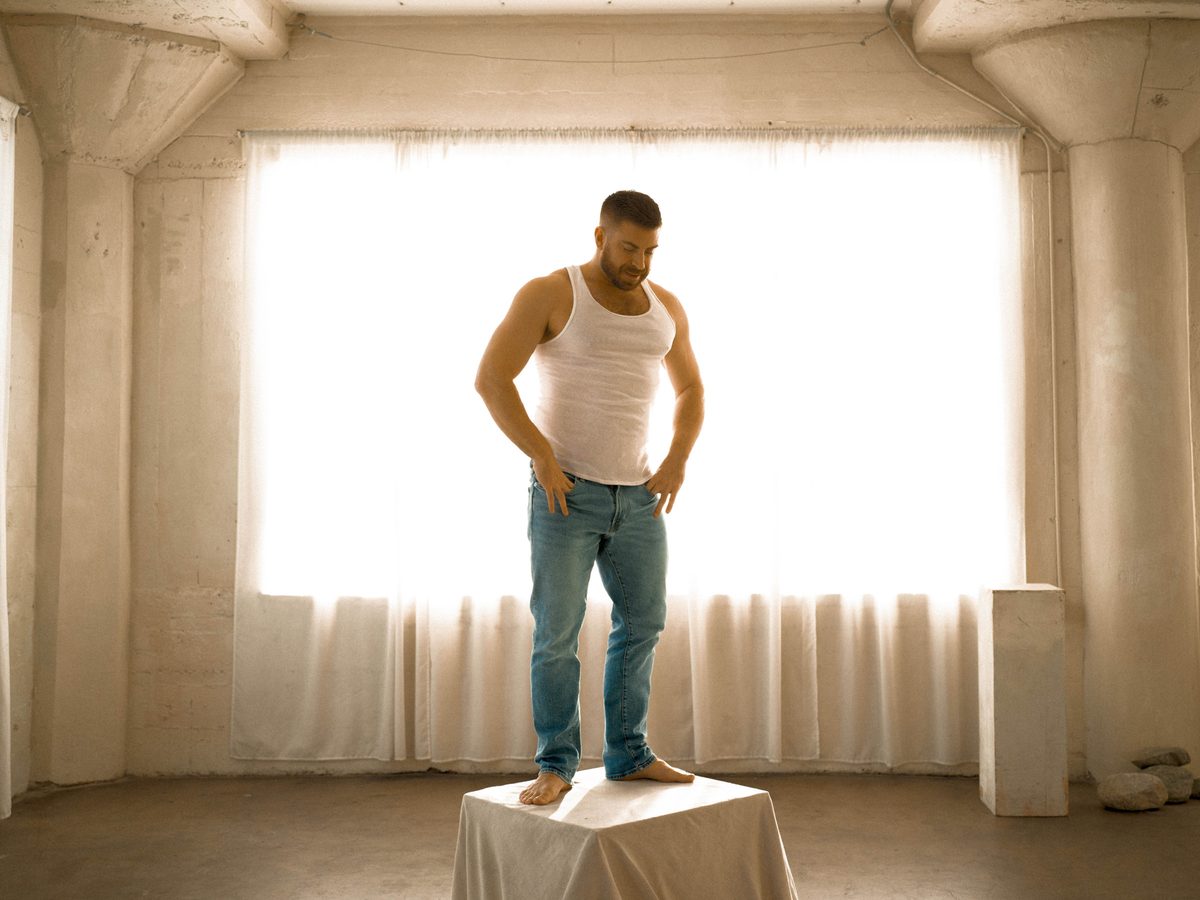Spread 1 of 10 from Studio Sessions: a bearded man in a tank top and jeans stands barefoot on a draped pedestal in a sunlit studio; colour.
