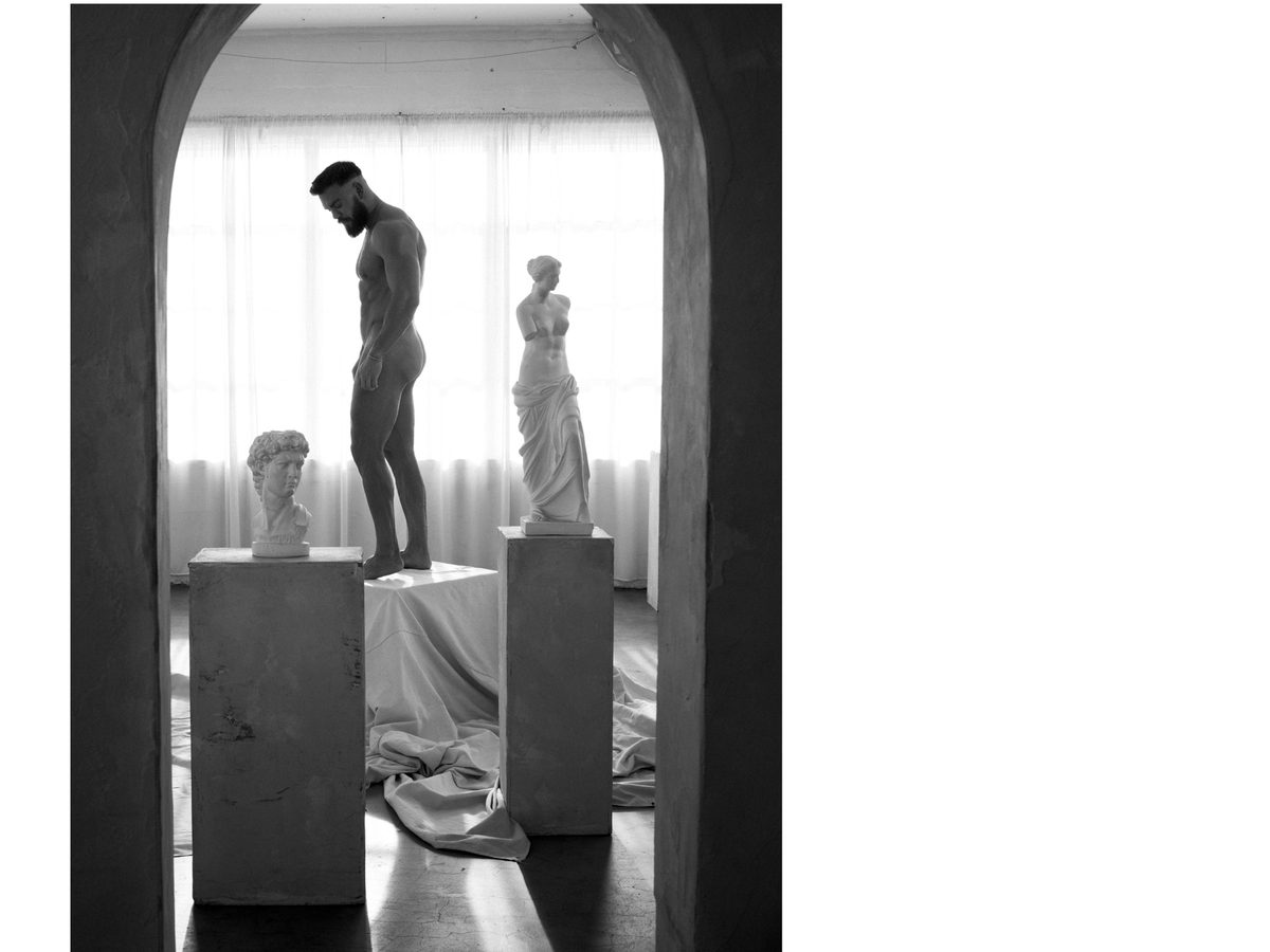 Spread 1 of 10 from Ancient Forms: a nude man stands on a pedestal beside classical plaster busts and statues in a sunlit studio; black and white.