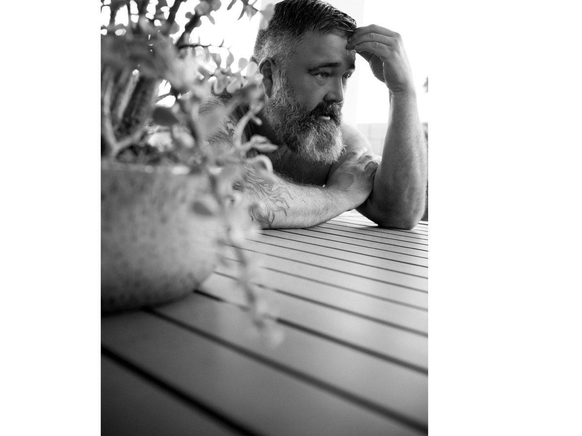 Spread 4 of 10: a bearded man leans on an outdoor table beside potted plants, looking aside; black and white.