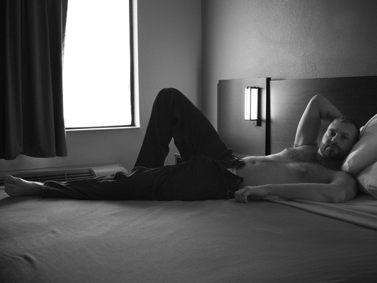 Spread 2 of 11 from Motel 6: a bearded, shirtless man lies across a motel bed in jeans, arm behind his head; black and white.
