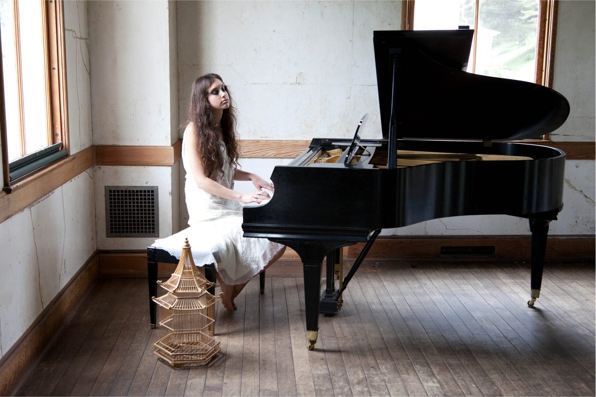 A woman in a pale dress kneels at the bench of a grand piano in a sparse, light-filled room; colour.