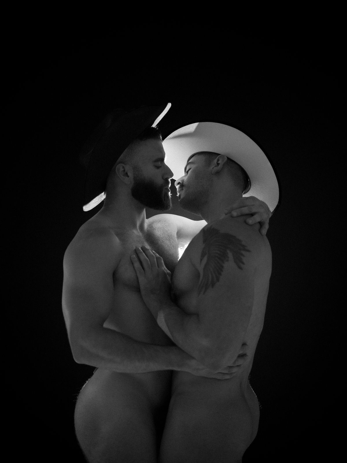Two nude men embrace face to face in a dark studio, rim-lit in profile, one with a winged tattoo on his back; black and white.
