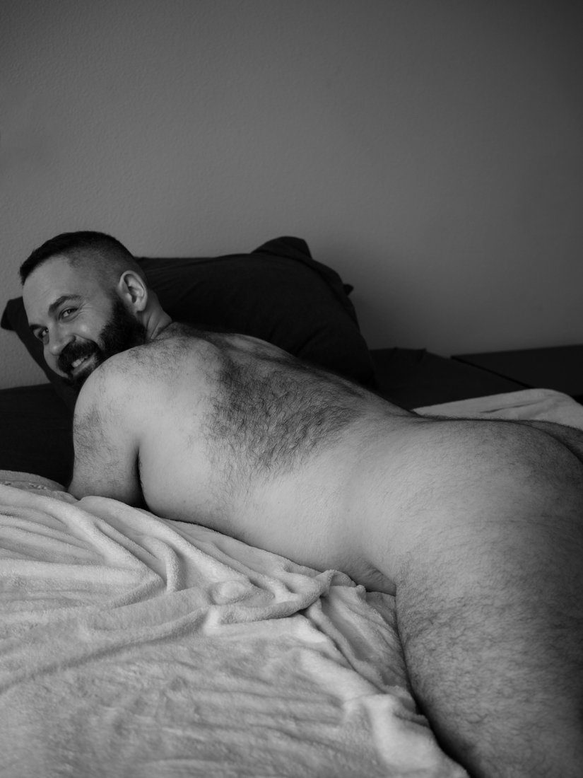 A bearded, heavyset man lies nude across a bed, propped on one arm, smiling toward the camera; black and white.