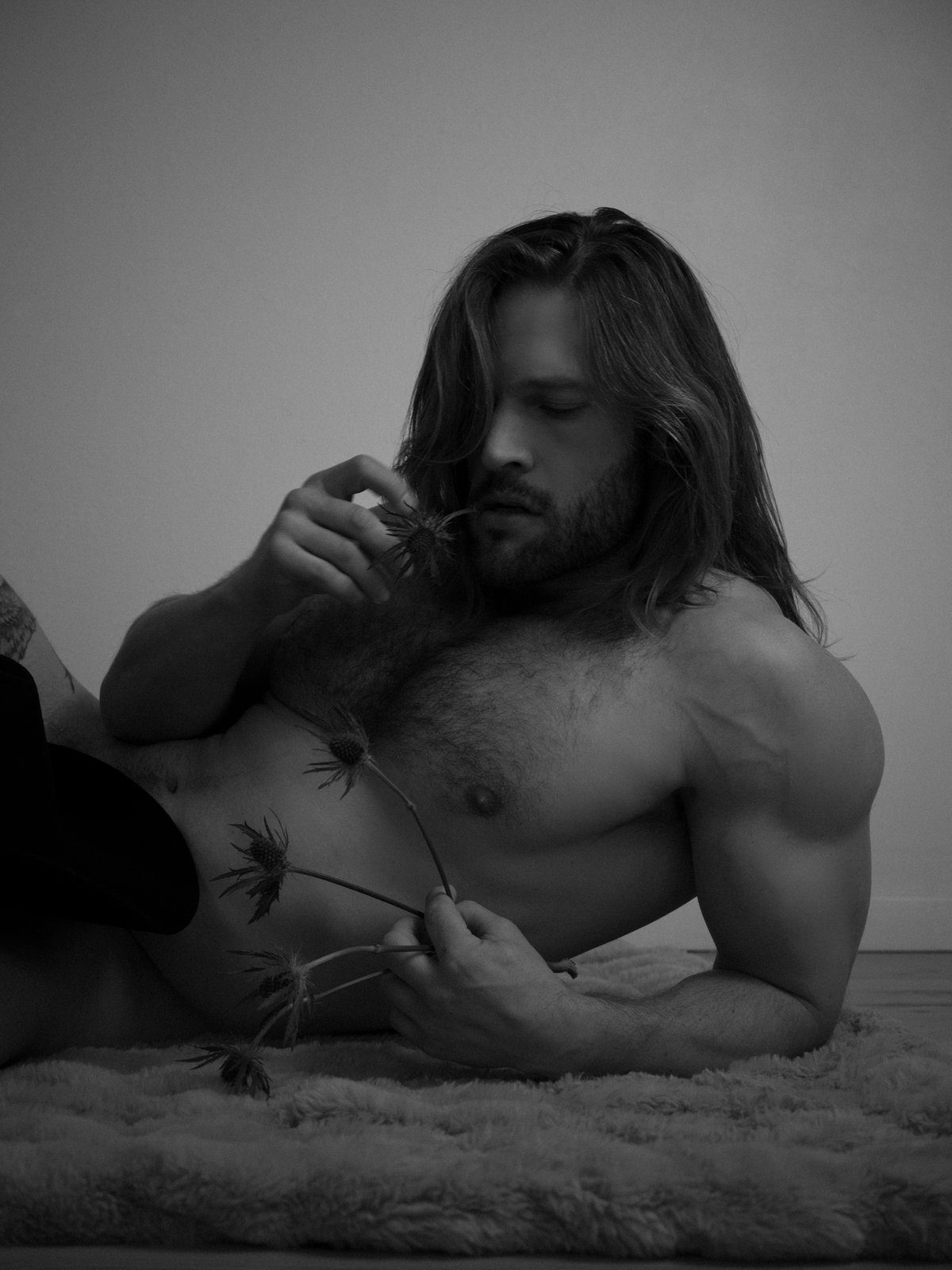 A long-haired, bearded, muscular man lies on a fur rug, shirtless, examining a dried flower stem in his fingers; black and white.