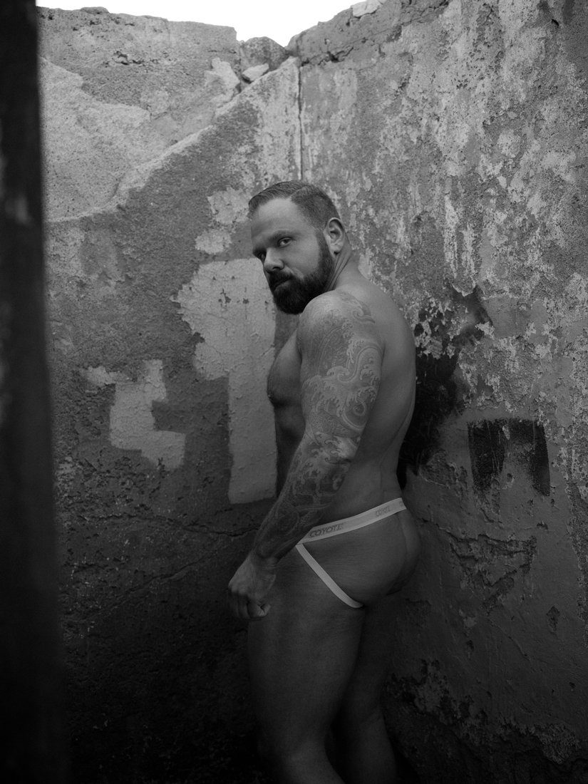A bearded, tattooed man leans against a weathered stucco wall in briefs, looking off to the side; black and white.