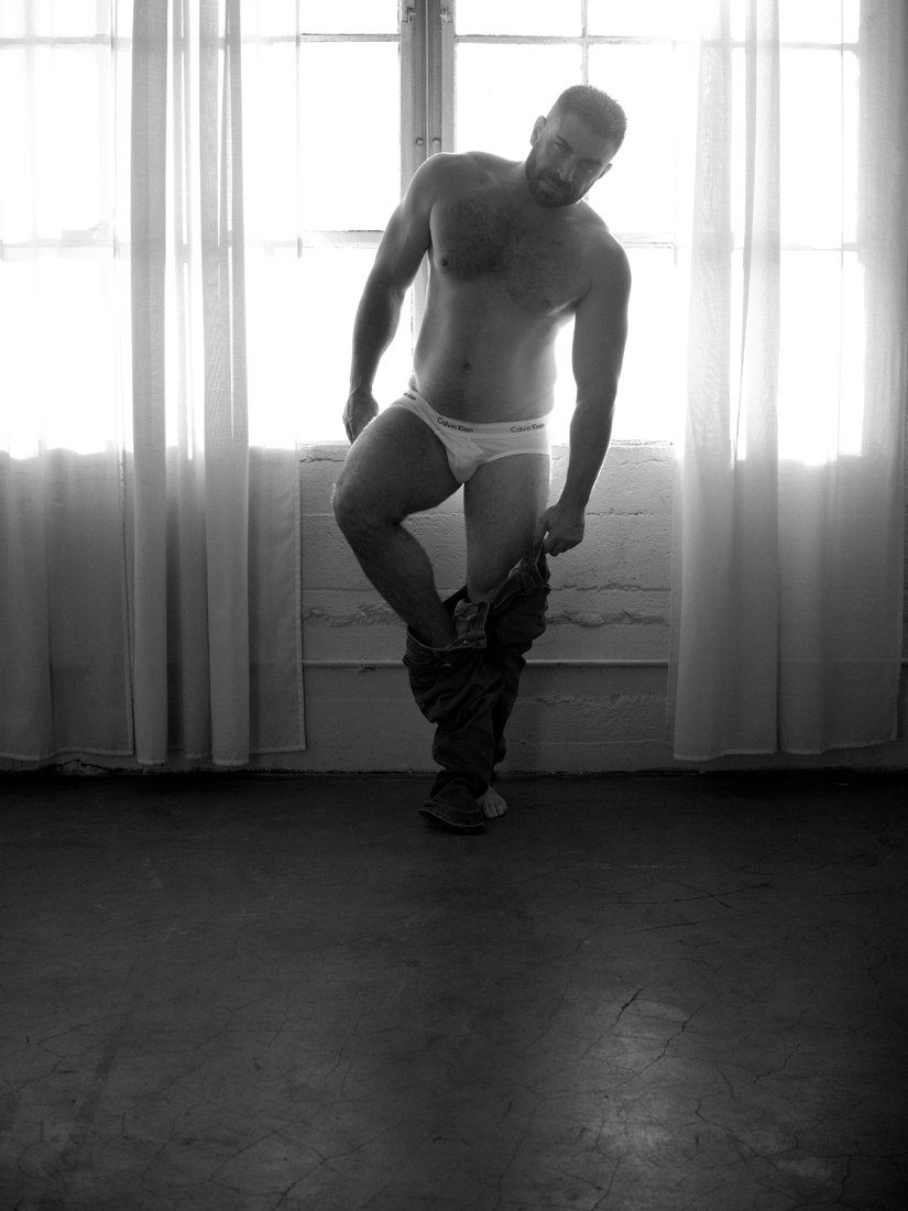 A bearded man in briefs balances on one leg by a sunlit window, removing his trousers; black and white.
