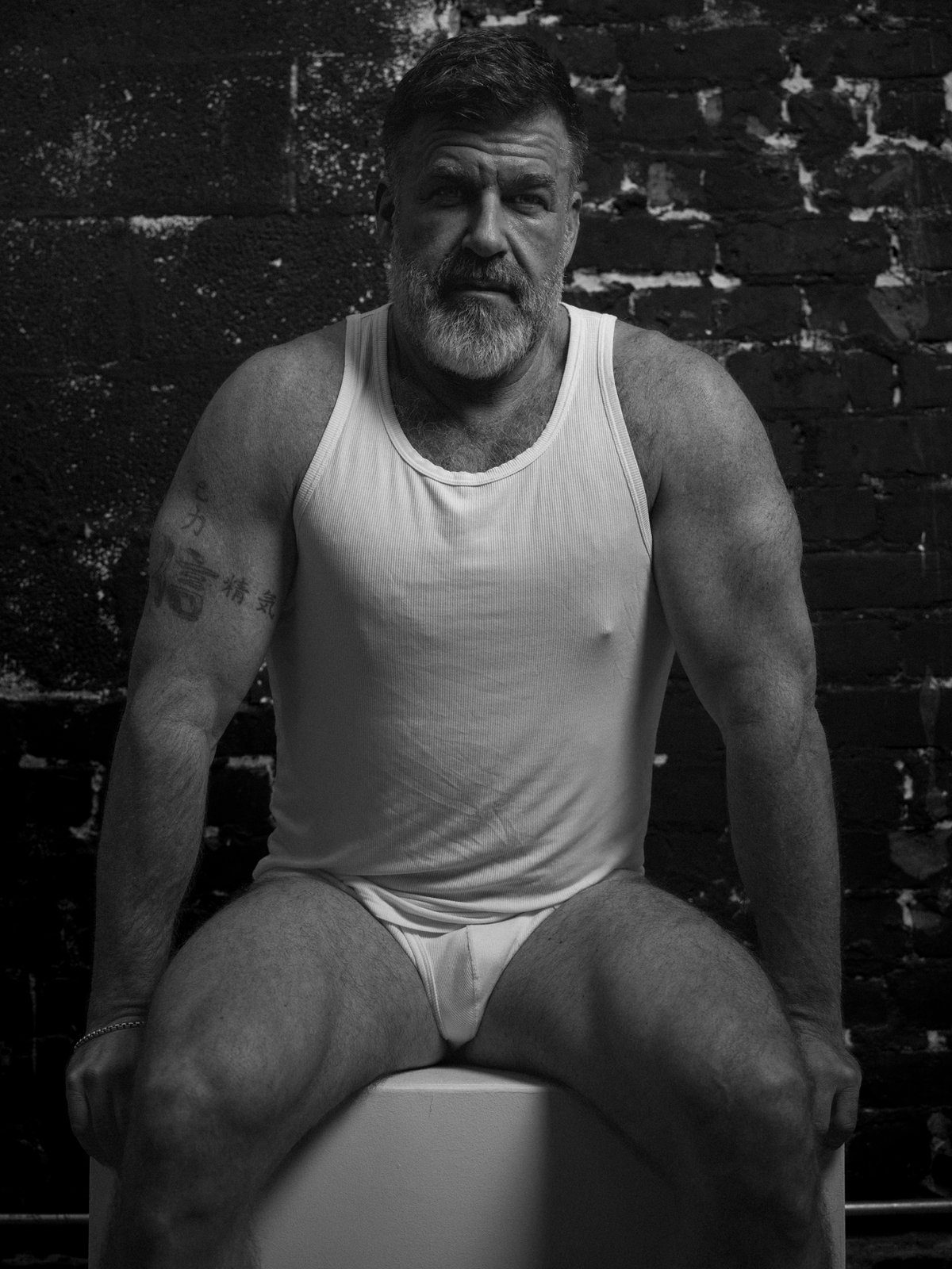 A bearded older man in a white tank top and briefs sits on a white block against a brick wall, looking at the camera; black and white.