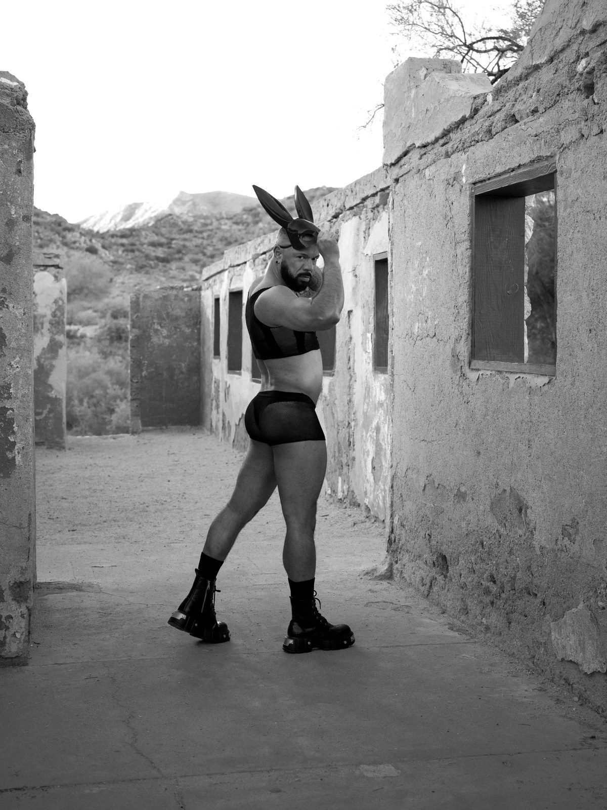 A man in a black harness, briefs and tall boots wears rabbit ears, standing in a ruined adobe passage with mountains behind; black and white.