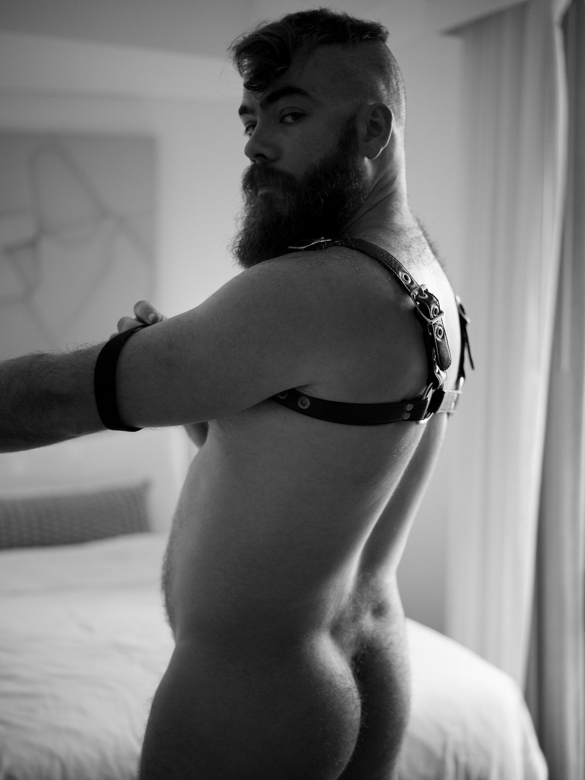 A bearded man in a black leather harness kneels on a bed, seen from behind, adjusting the strap at his shoulder; black and white.