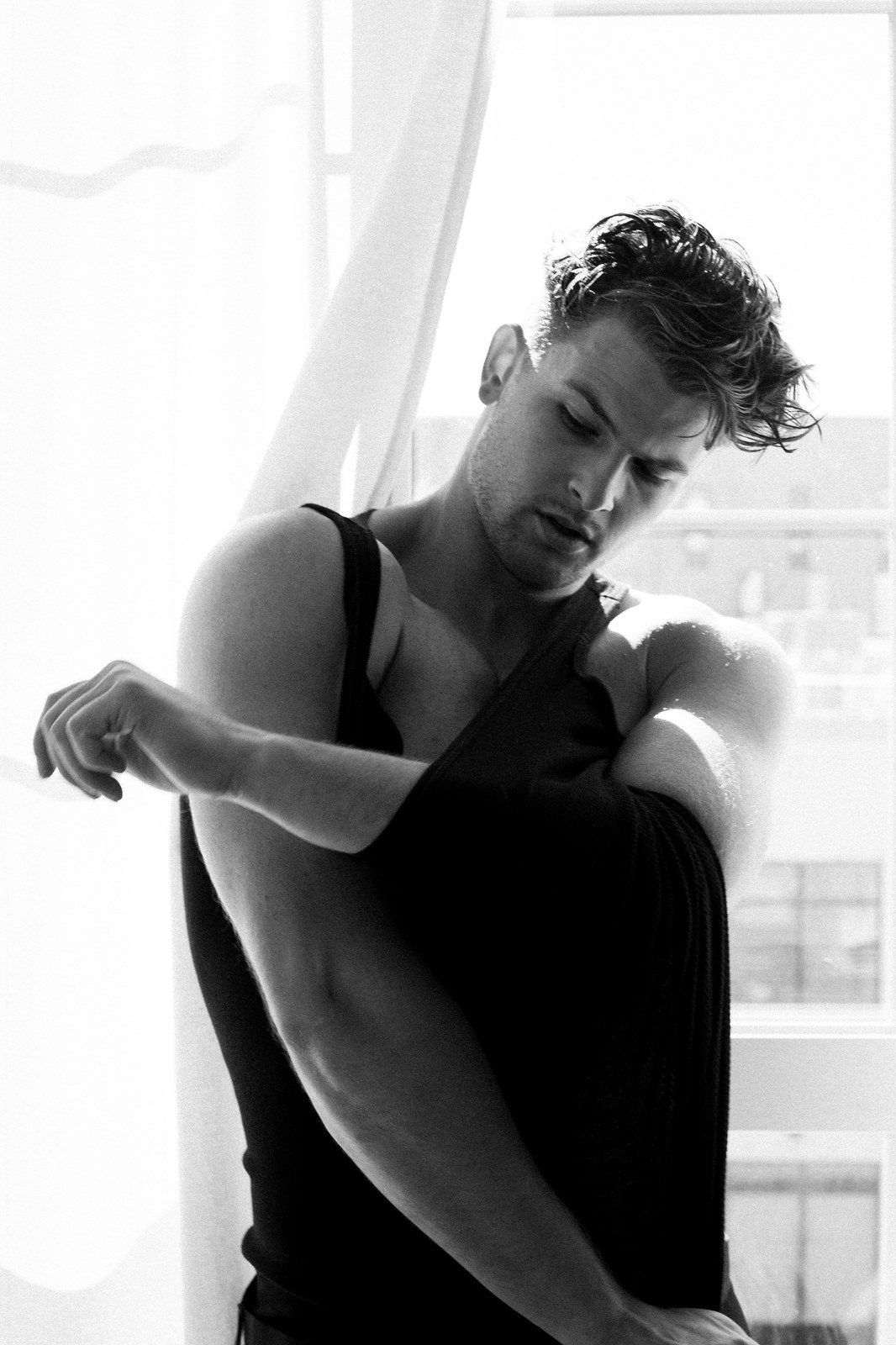 A man in a black tank top stands by a window, pulling the strap over his shoulder, hair damp; black and white.