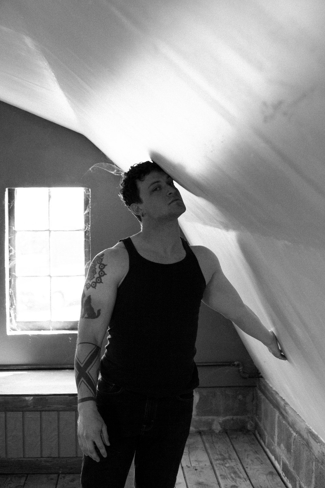 A tattooed man in a black tank top leans against draped fabric by a window, looking up; black and white.