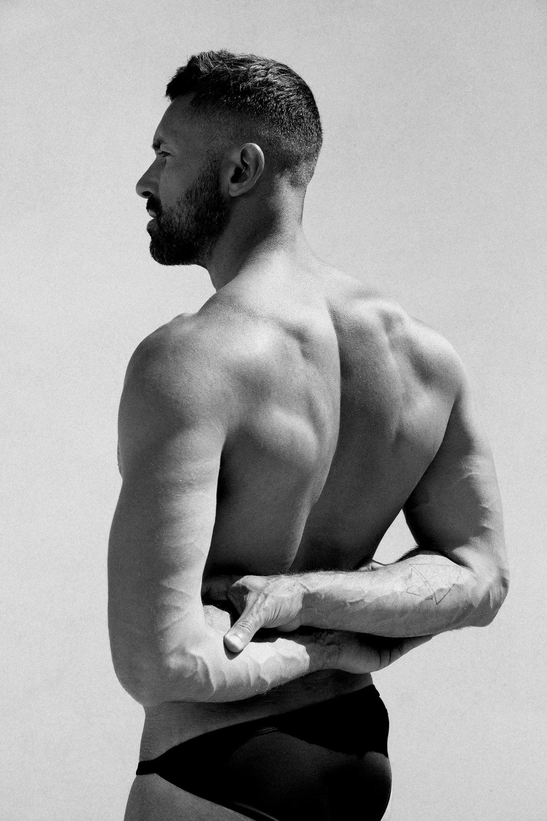 A bearded man stands in profile wearing dark briefs, arms crossed behind his back, studio light; black and white.
