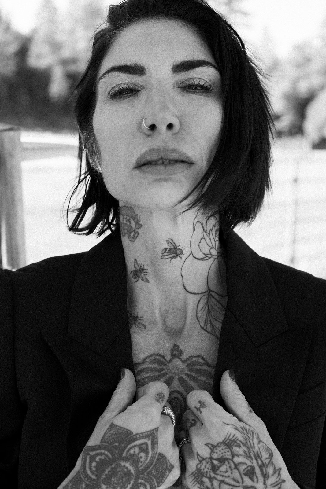 A woman in a black blazer with a tattooed chest and neck holds her hands together at her chest; black and white.